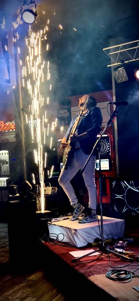 Musician playing guitar on stage with sparks and lights, stage equipment visible, dark background, colorful lighting.