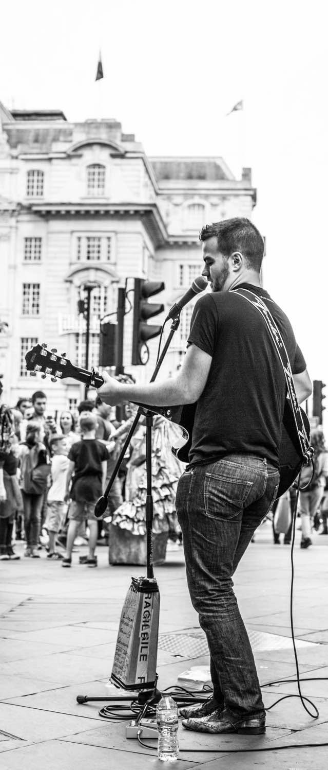 Street performer playing guitar and singing in a busy outdoor area with people and buildings in background.