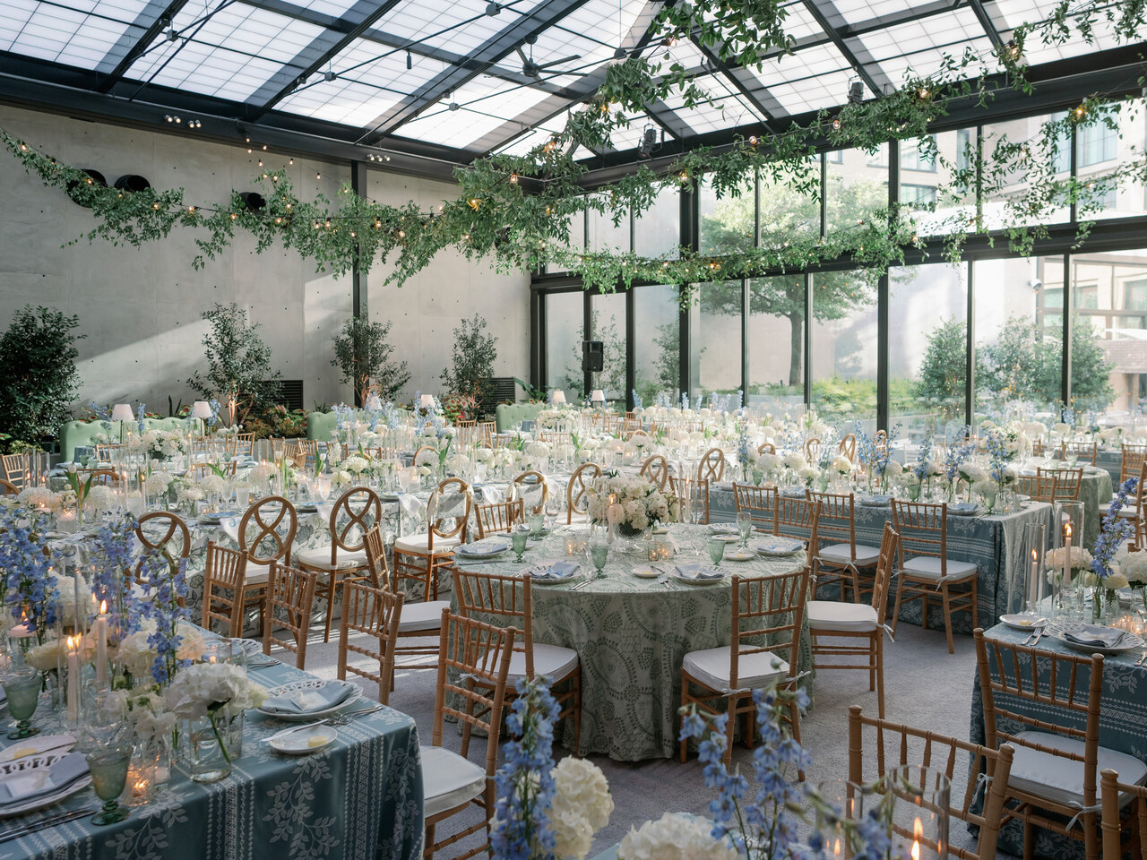 Elegant indoor event space with large glass ceiling, decorated tables with floral centerpieces, and greenery hanging from ceiling.