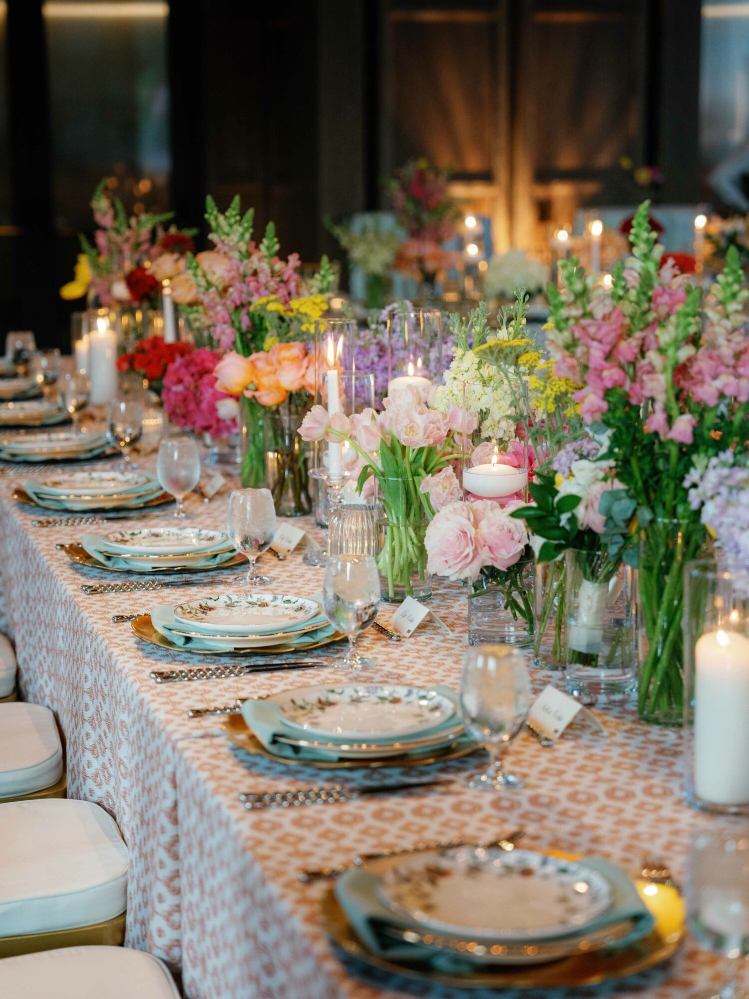 Long table decorated with colorful flowers, candles, and elegant tableware in a warmly lit room.