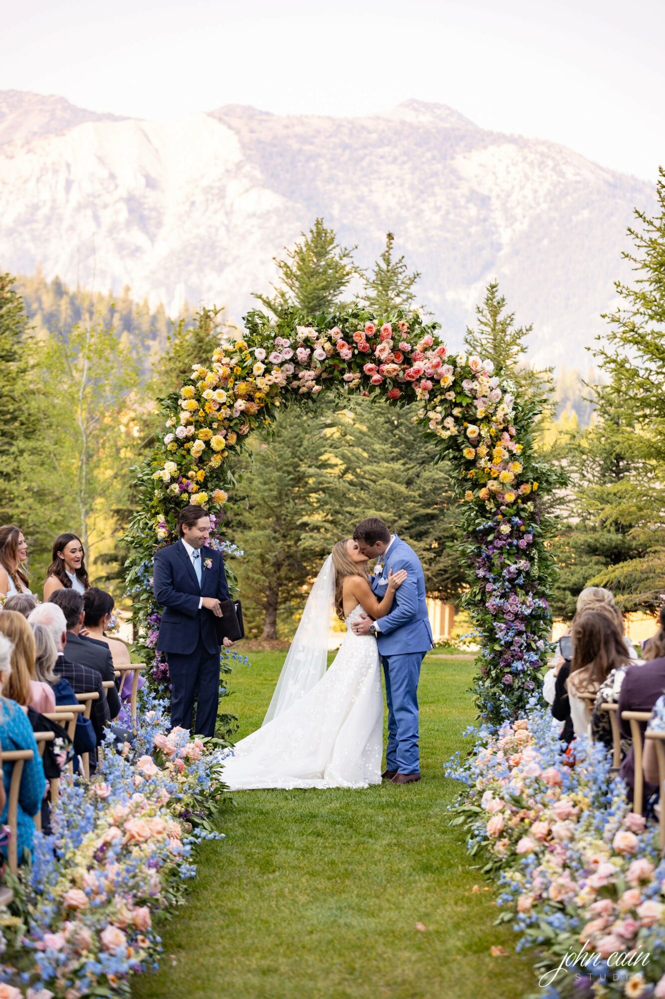 Couple kissing under a floral arch at outdoor wedding ceremony with guests and mountains in background.