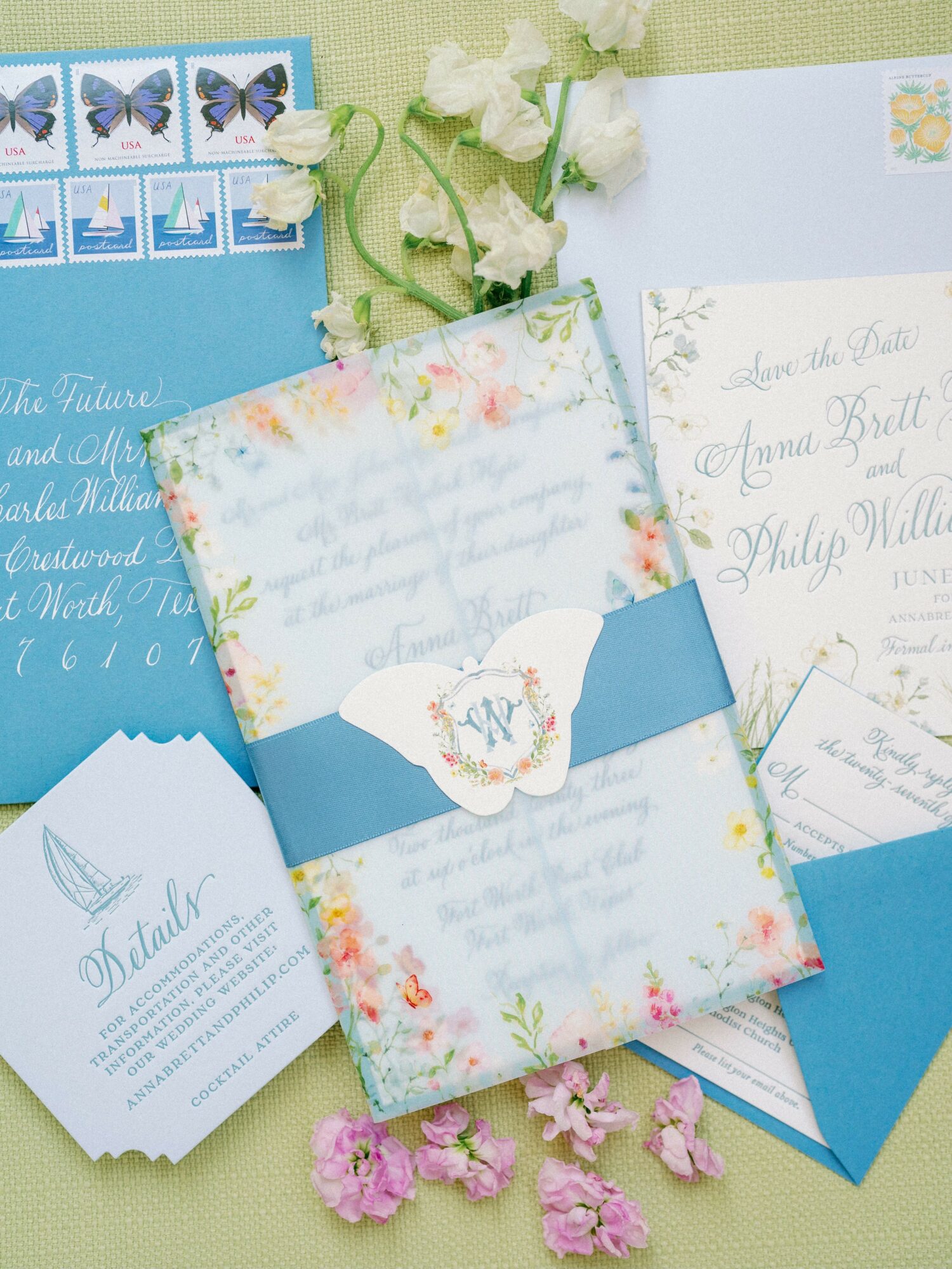 Wedding invitation with floral design, blue accents, and handwritten calligraphy, surrounded by flowers and stationery on a green background.