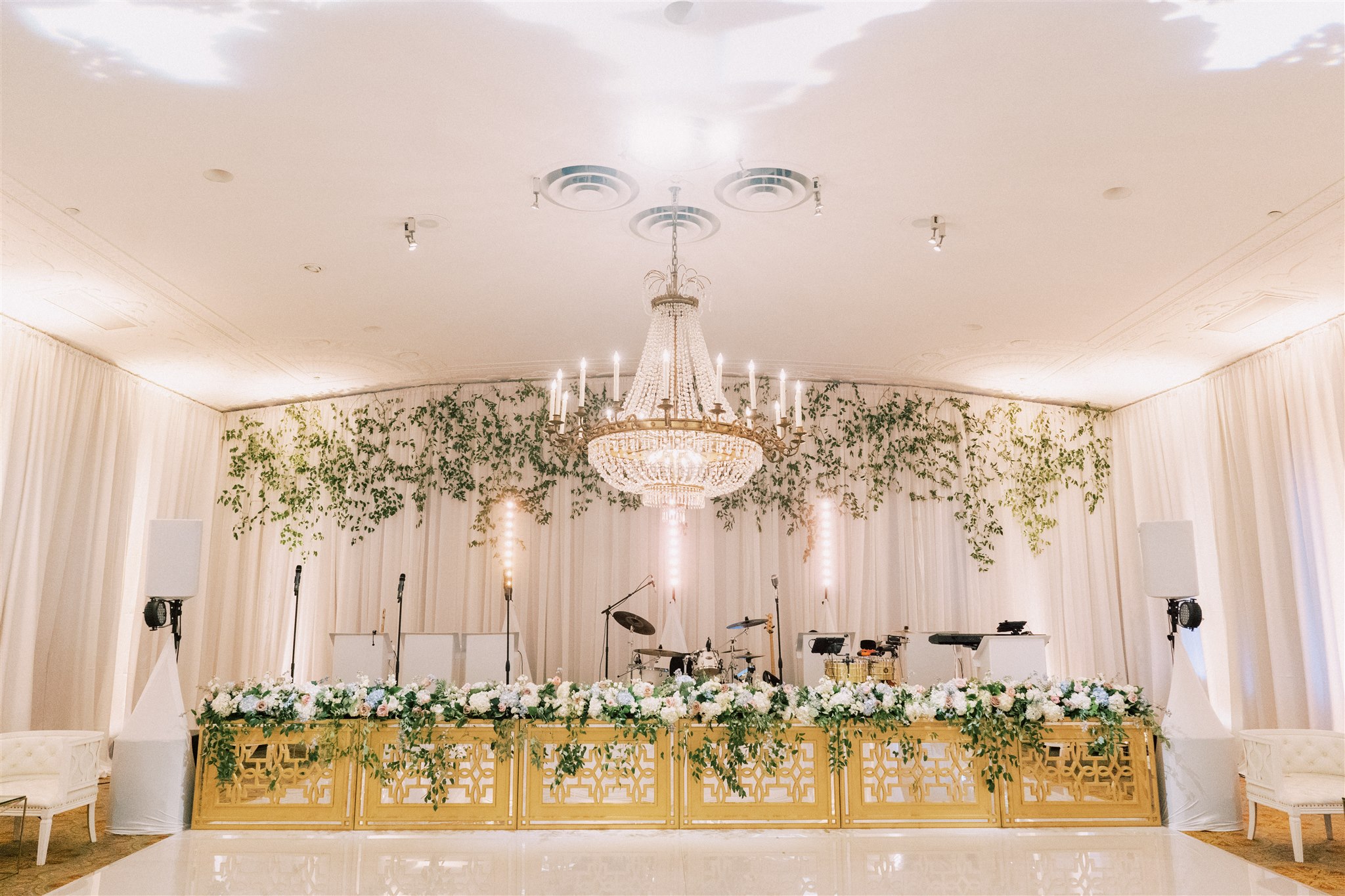Elegant event stage with floral decorations, chandeliers, and musical instruments, set in a bright, decorated room with white curtains.