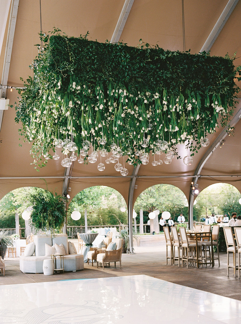 Hanging floral arrangement with green foliage and white flowers inside a decorated event space with tables and chairs.