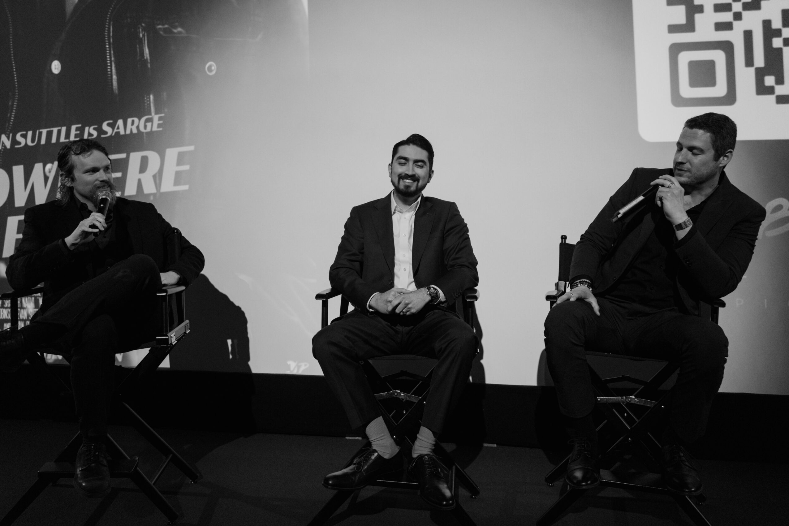 Three men sit on stage in chairs, engaged in a discussion, with a large screen behind them displaying text and a QR code.