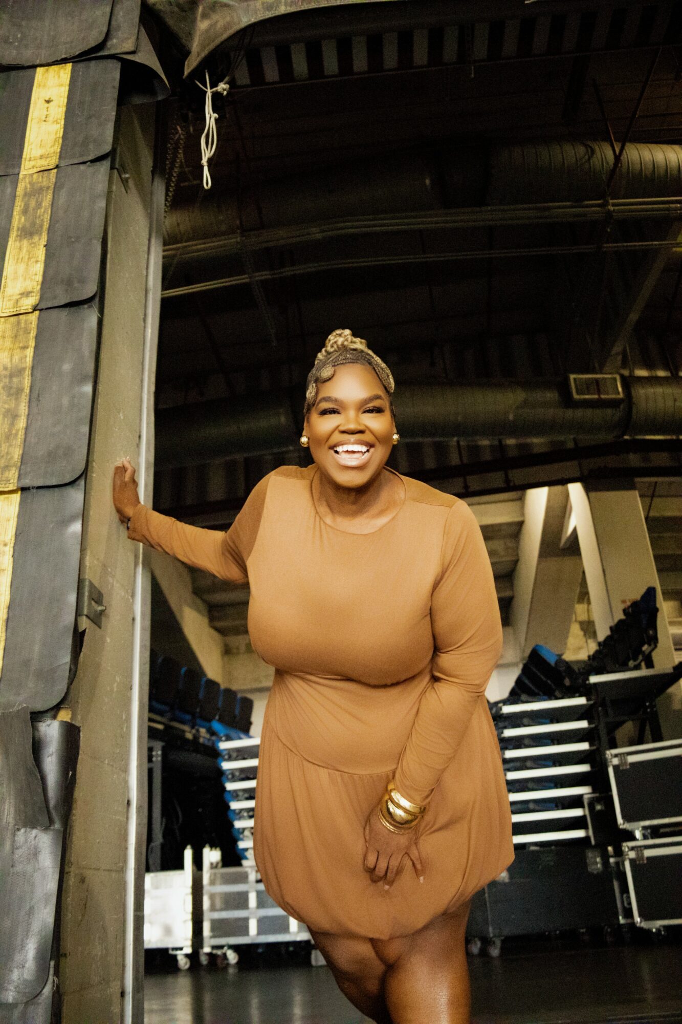 Smiling woman in a tan dress standing in an industrial setting, holding a small object, with a large metal structure behind her.
