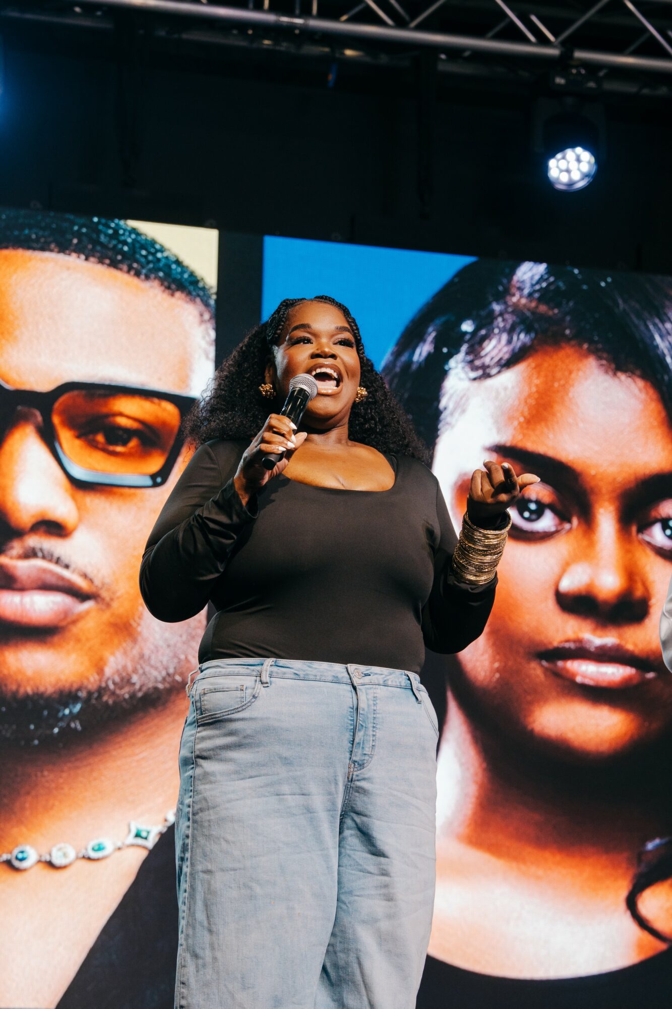 Woman speaking into microphone on stage with large portraits of two people behind her.