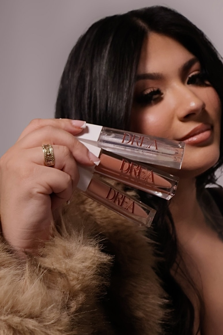 Woman with dark hair holding three lip gloss tubes, wearing a fur coat, looking at the camera.