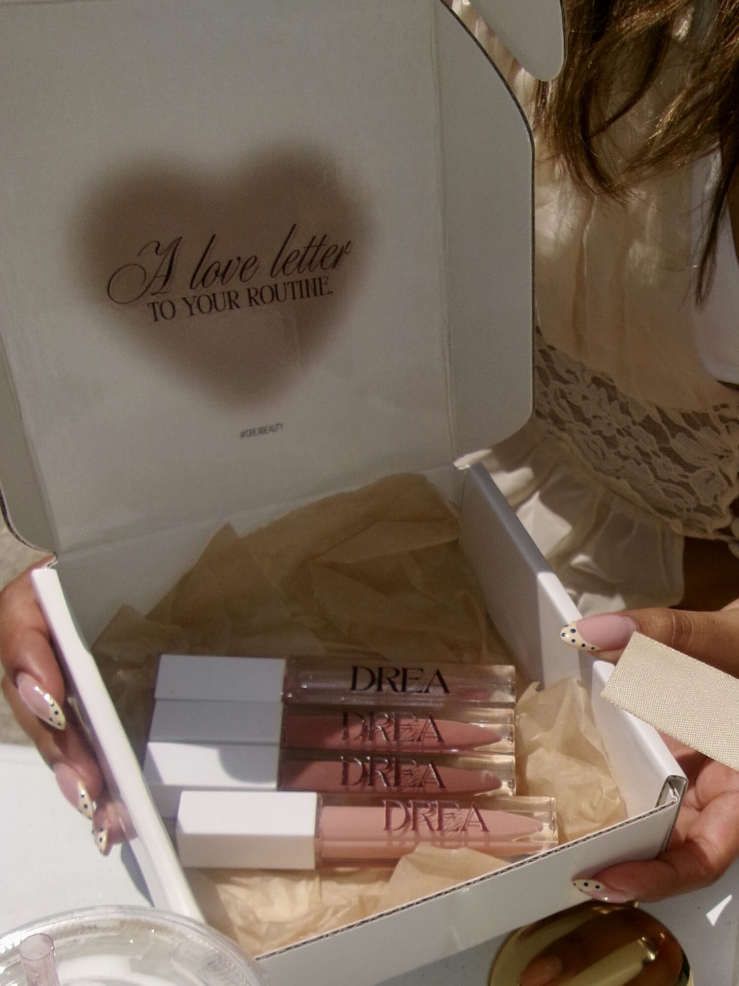 Open box with four lip glosses inside, heart-shaped message on lid, person partially visible on right, beige tissue paper inside box.
