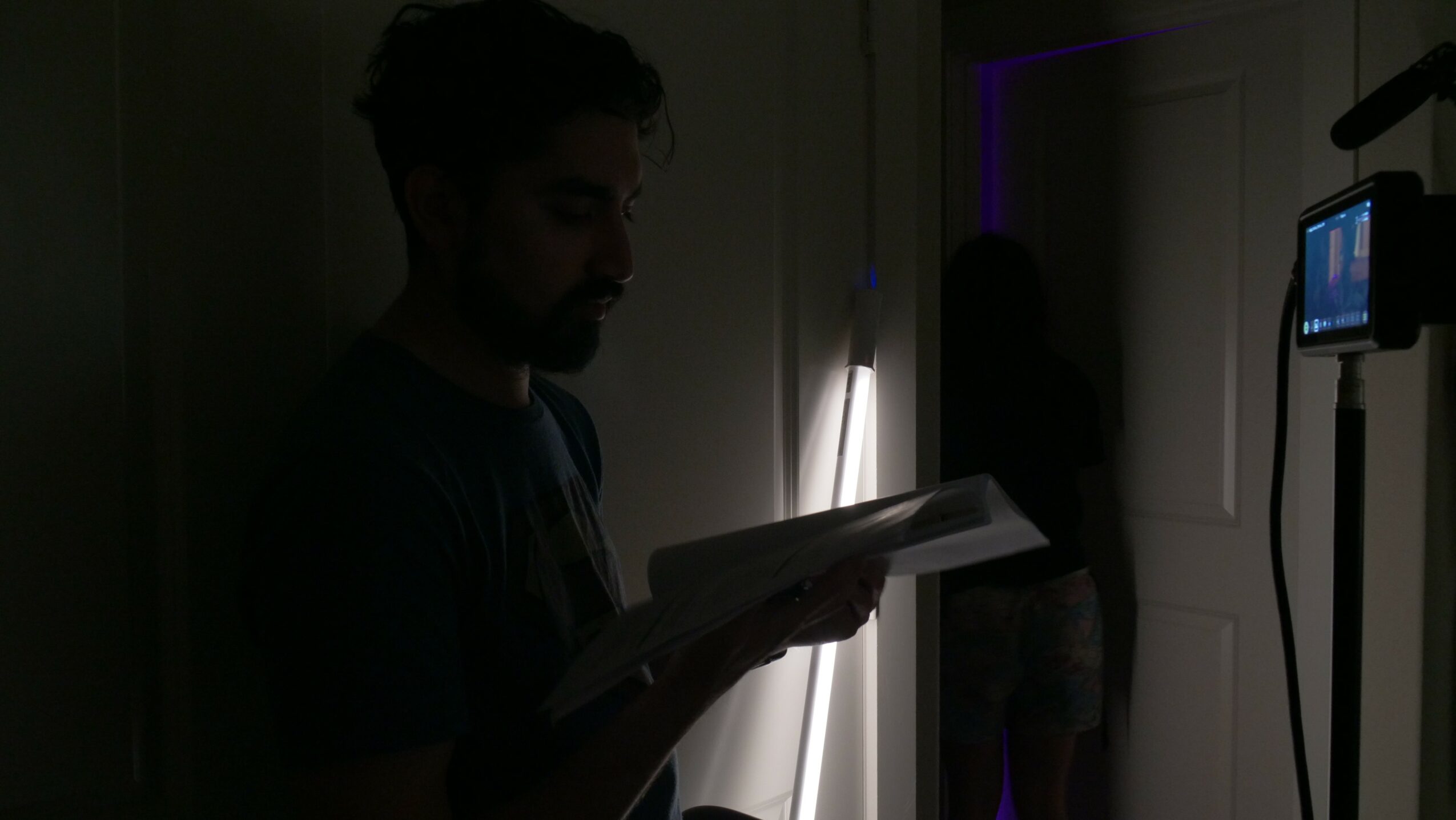 Person reading a book in a dimly lit room with a door and a person in the background.