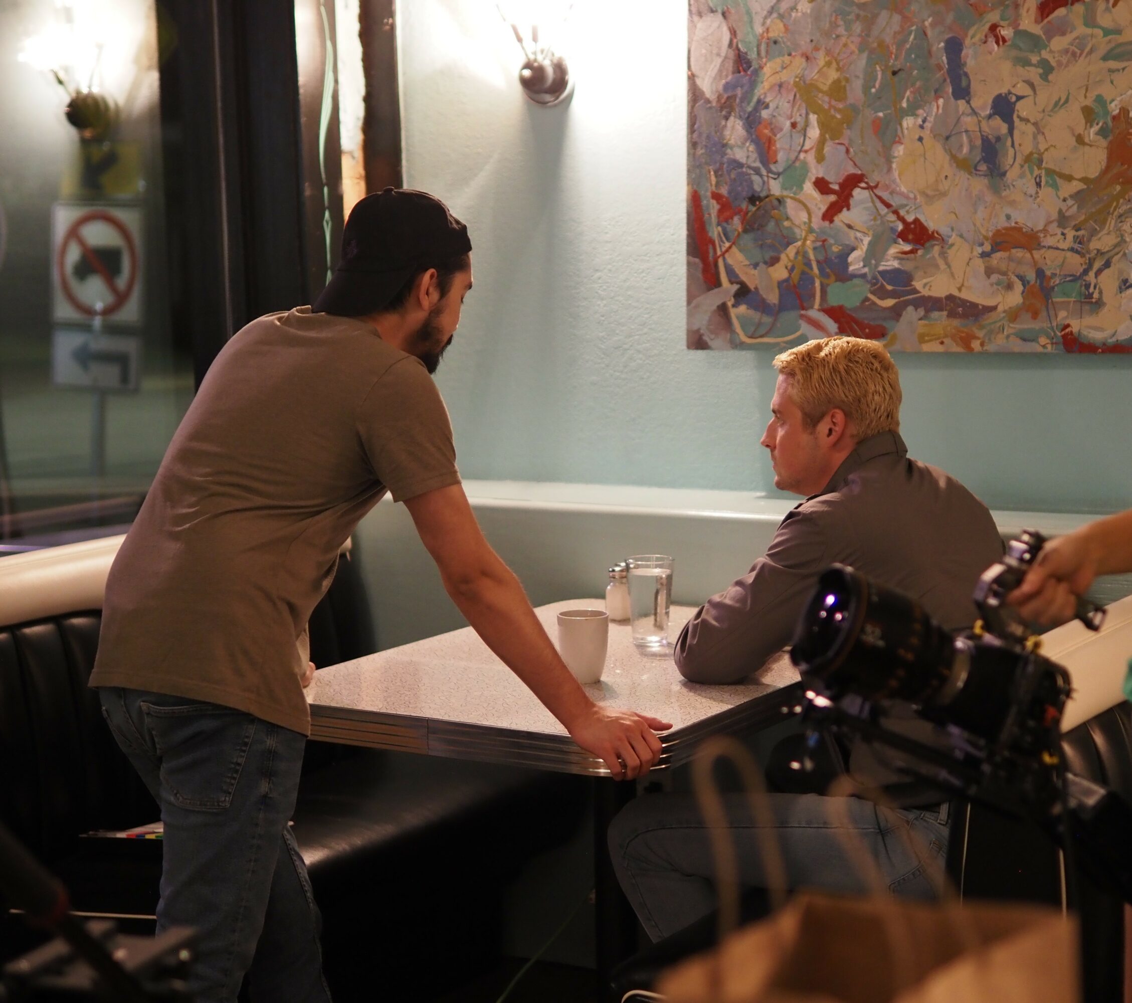 Two men in a restaurant, one standing and leaning on the table, the other sitting and looking at him, with a colorful abstract painting on the wall.