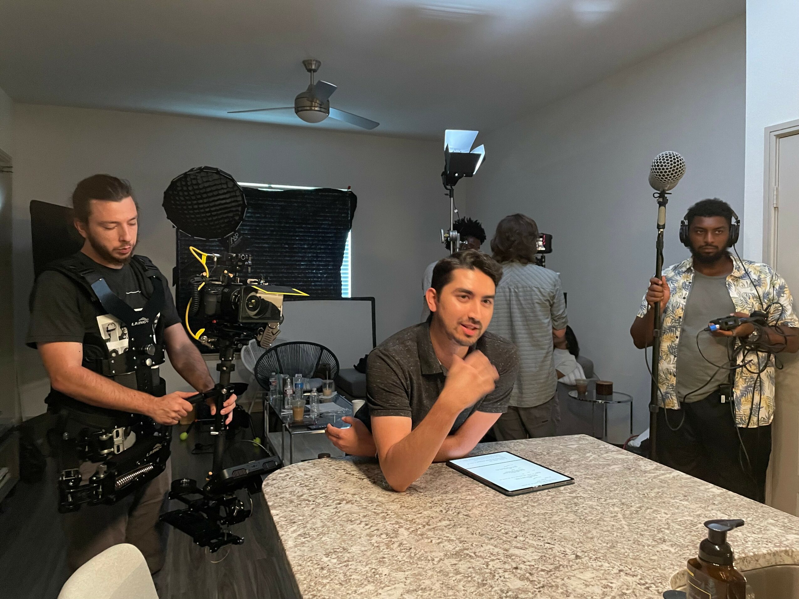 Man sitting at a table with a tablet, surrounded by camera crew and lighting equipment in a room.
