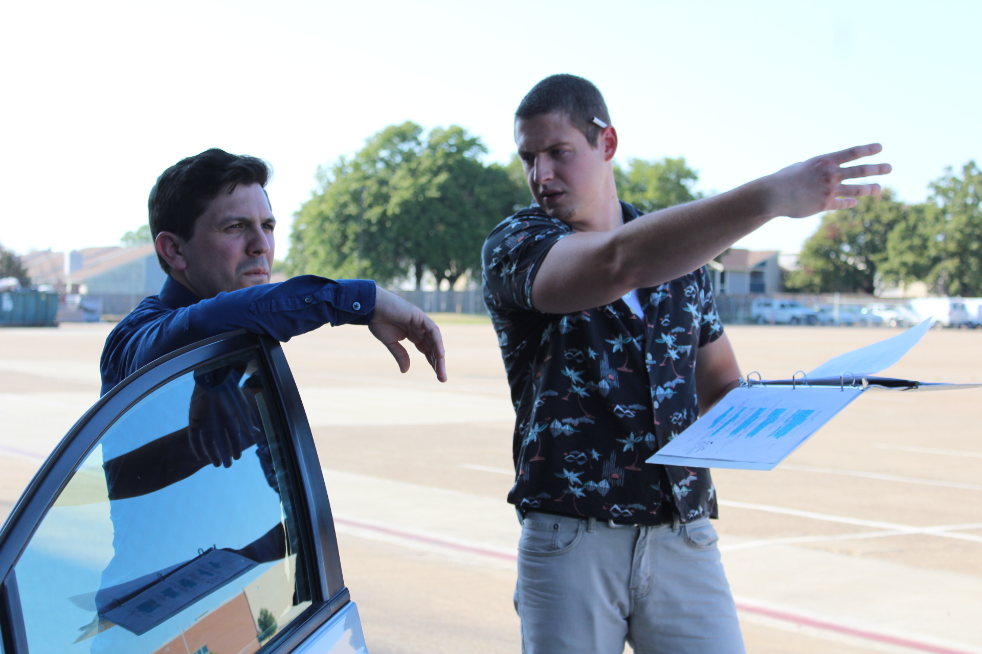 Two young men outdoors, one pointing and the other leaning on a car, with trees and a field in the background.