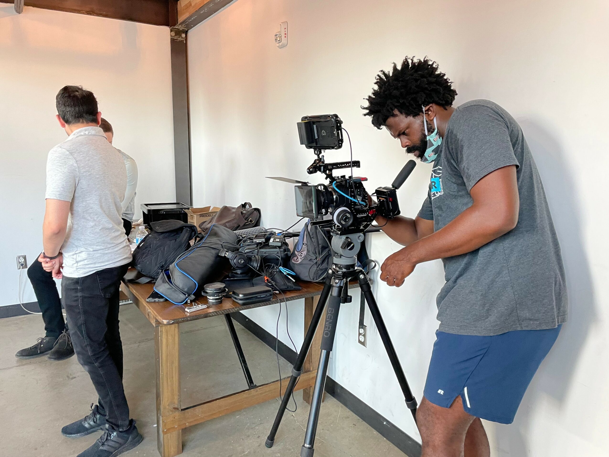 Person with curly hair operating a camera on a tripod, adjusting settings, in a room with a table and bags, two other people nearby.