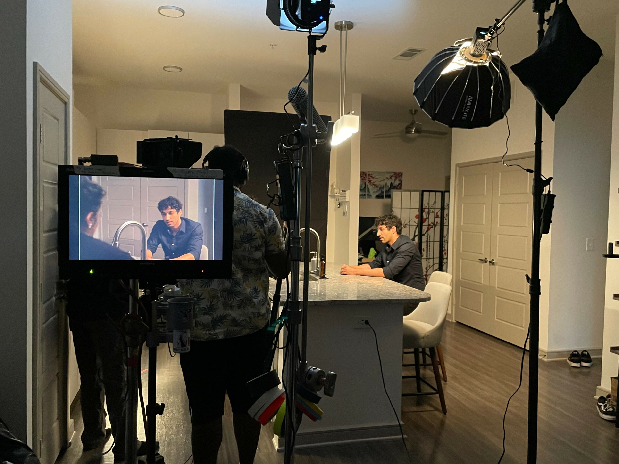 Person filming a man sitting at a kitchen counter with studio lighting and equipment, in a home setting.