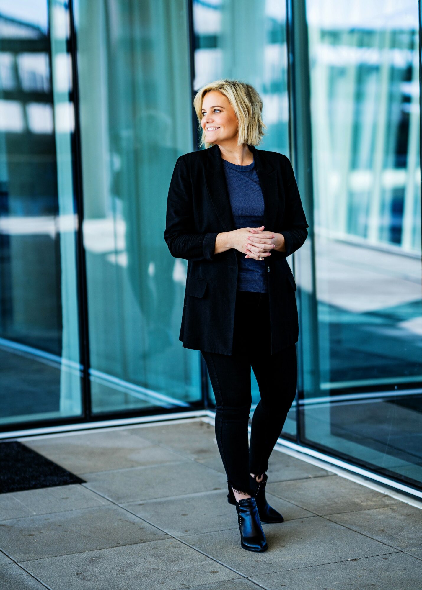 Woman with blonde hair in a black blazer and pants standing indoors near large glass windows, smiling and looking to the side.