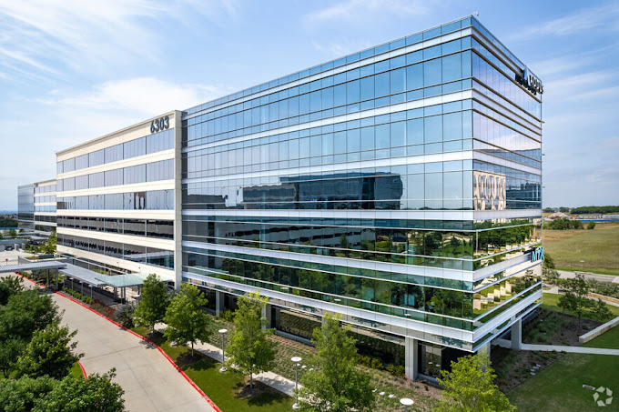 Modern glass office building with multiple floors, surrounded by trees and a parking lot, under a blue sky.