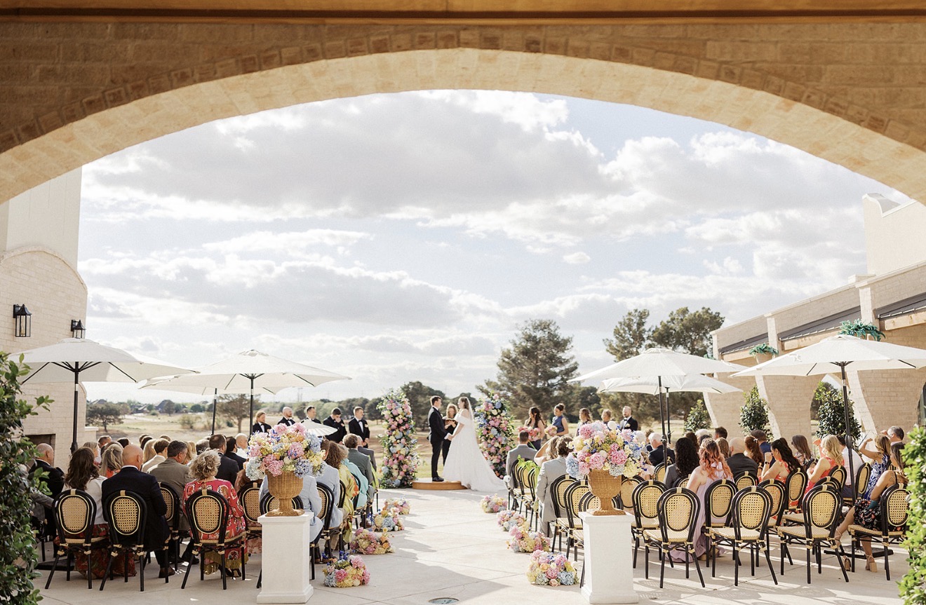 Outdoor wedding ceremony with guests seated under umbrellas, facing an arch, with a cloudy sky and trees in the background.