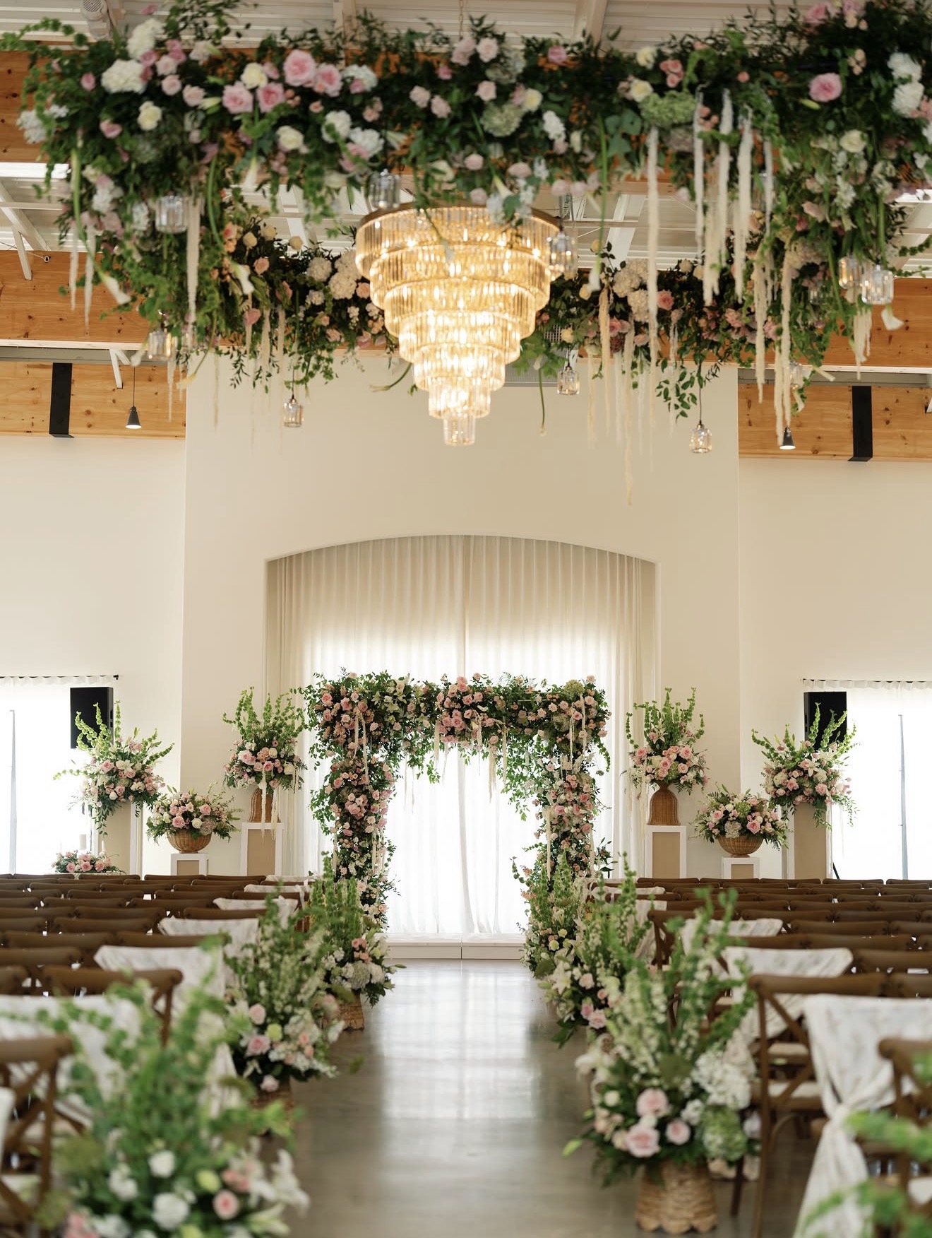 Elegant wedding ceremony setup with floral arch, aisle decorations, and chandelier in a bright, spacious hall.
