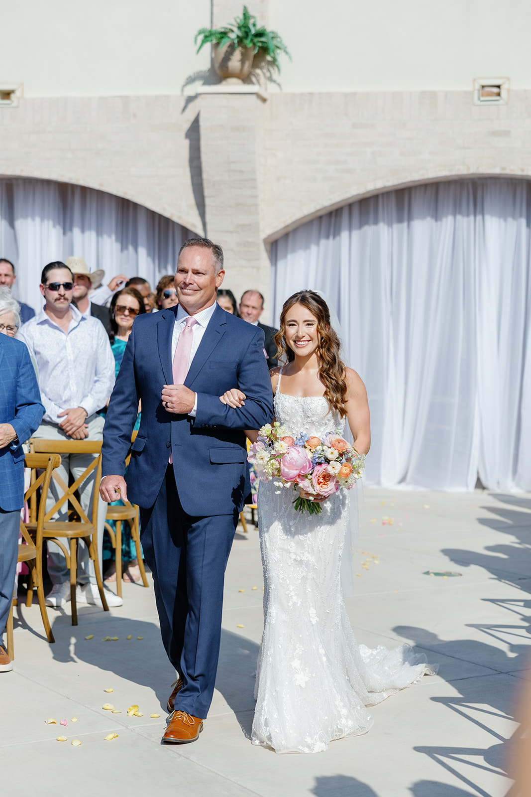 Bride in a white wedding dress holding a bouquet walking with a man in a suit outdoors, guests in background, sunny day.