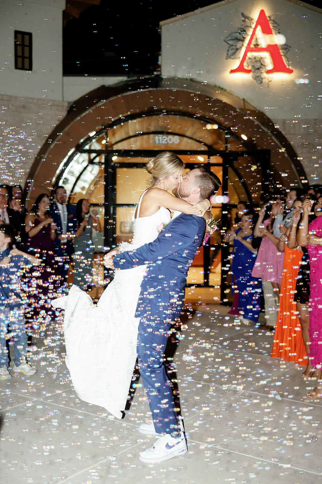 Couple dancing surrounded by guests, confetti falling, illuminated building with a large red letter A, nighttime scene.