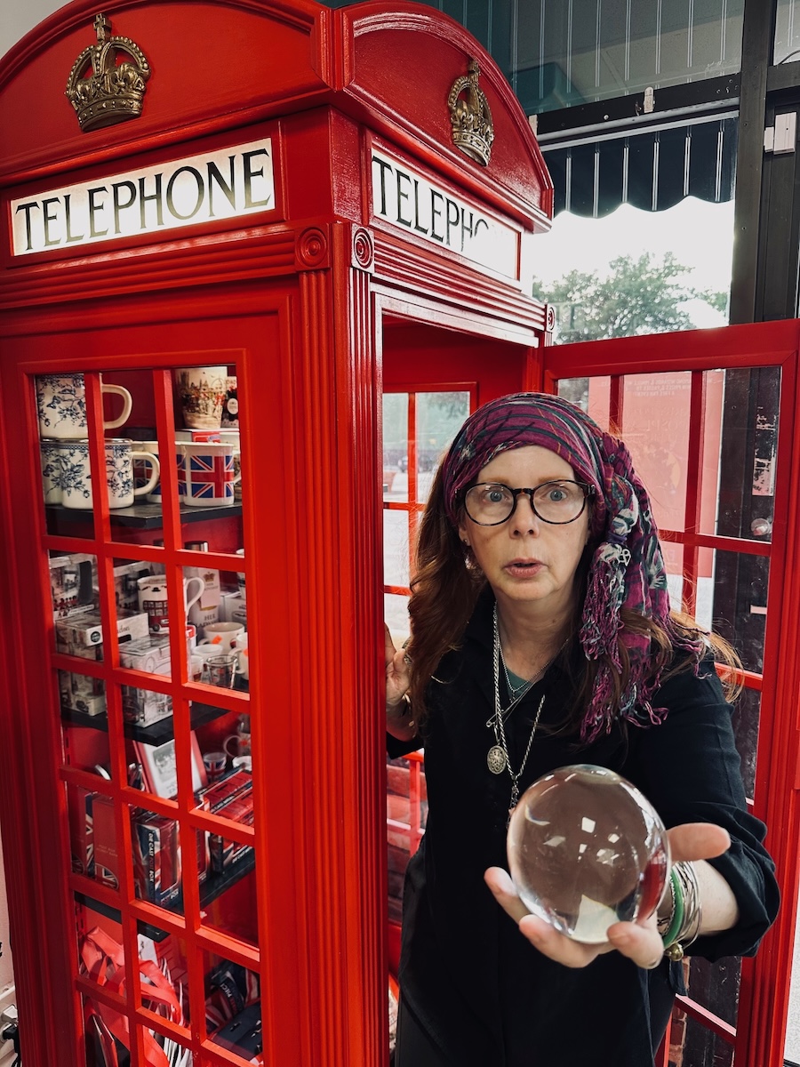 Woman with glasses holding a crystal ball inside a red telephone booth, with a window and outdoor scene in background.