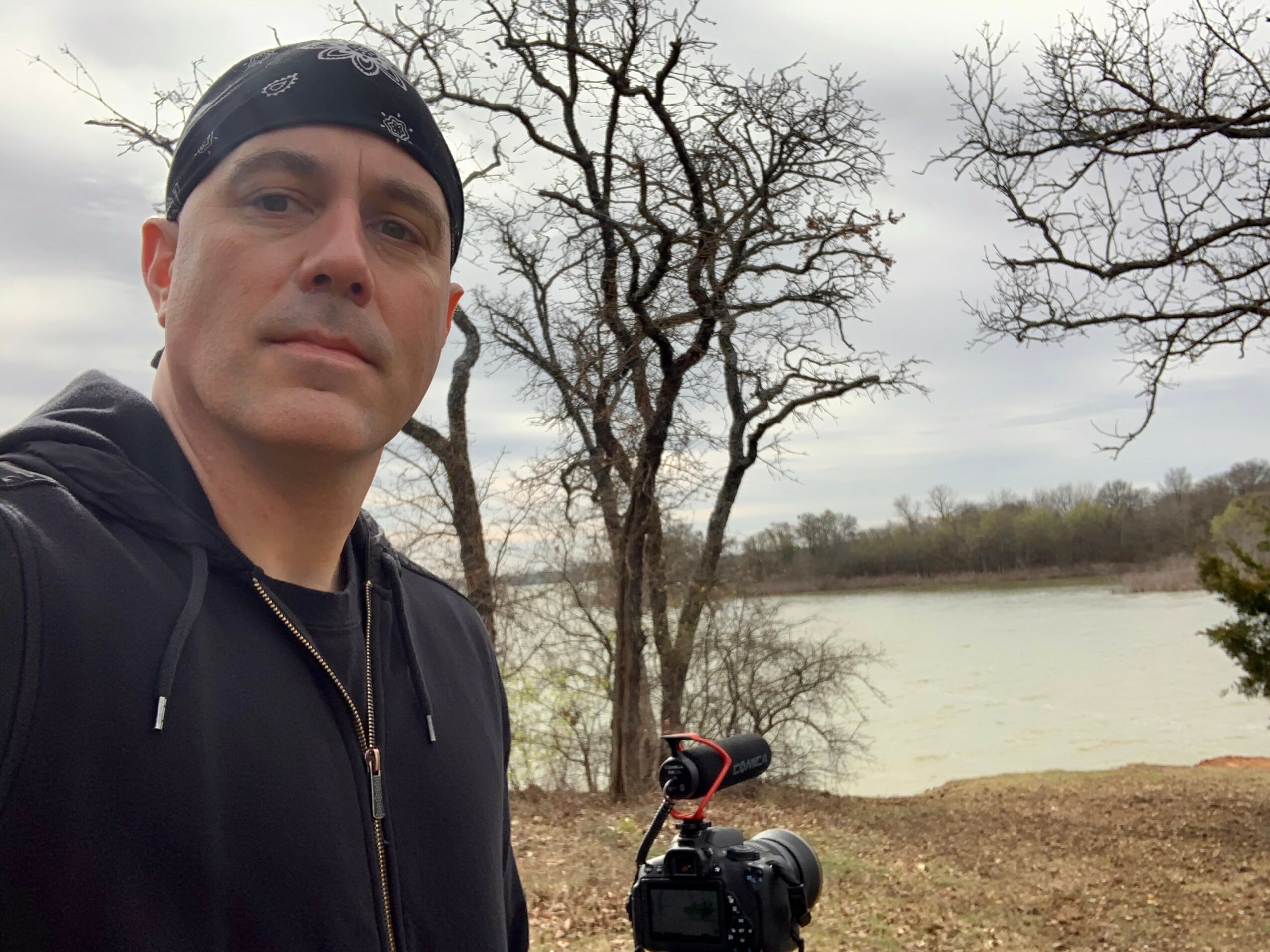Man wearing a black hoodie and headband outdoors near a river with leafless trees.