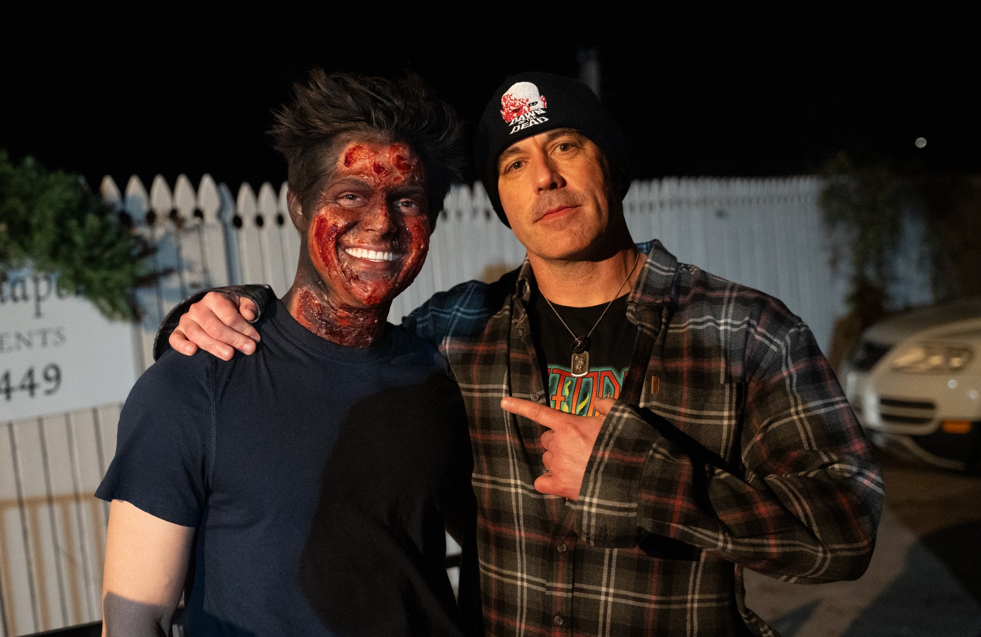 Two men standing outdoors at night, one smiling with face paint, the other pointing at him, both wearing casual clothing.