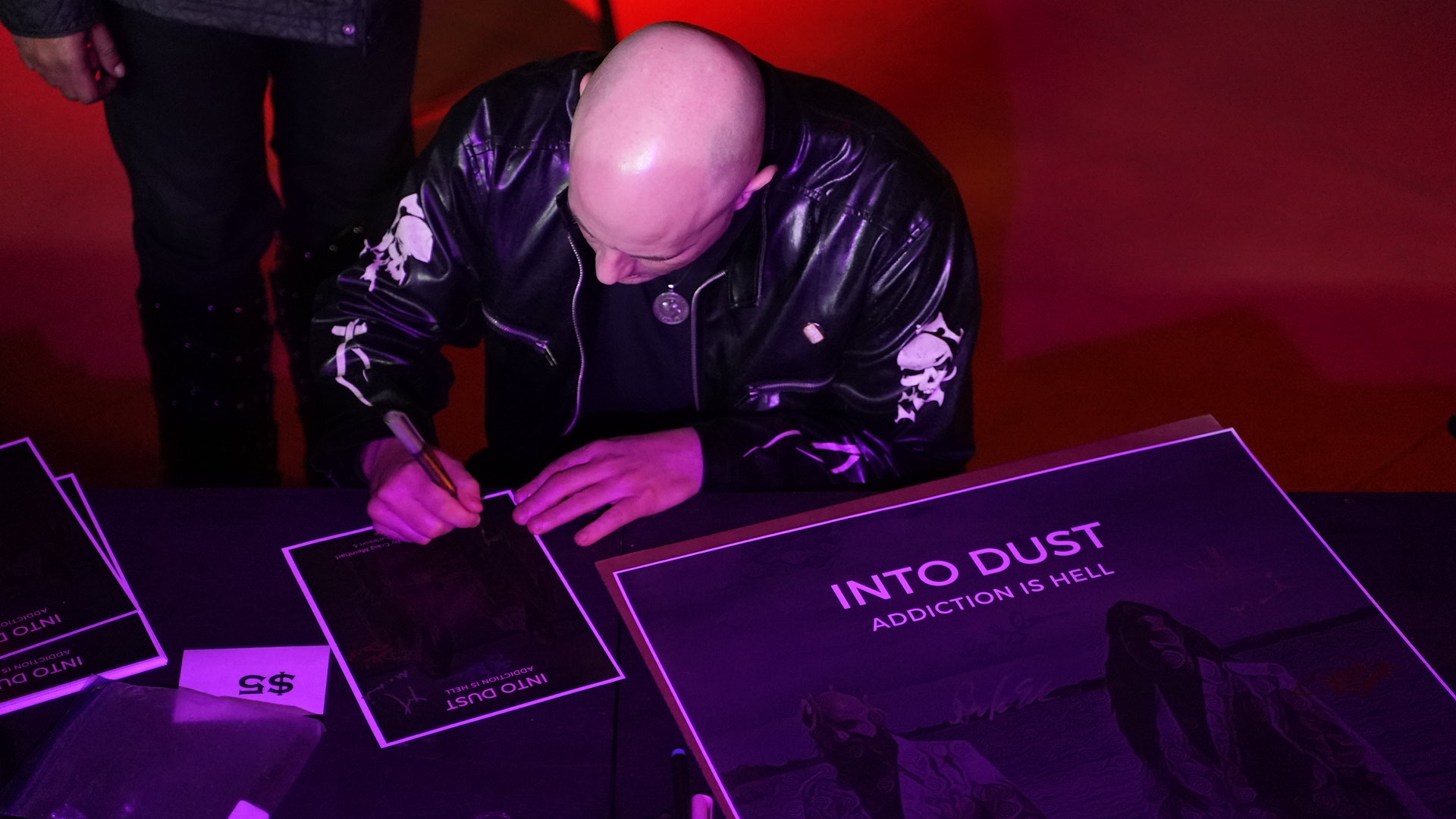 Bald person in a black jacket signing a book at a table with purple lighting, with a large sign in front.