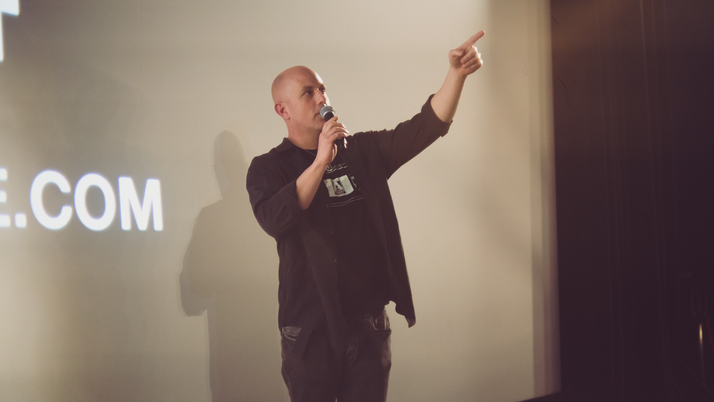 Person speaking into microphone, pointing to the right, on stage with a screen behind them.