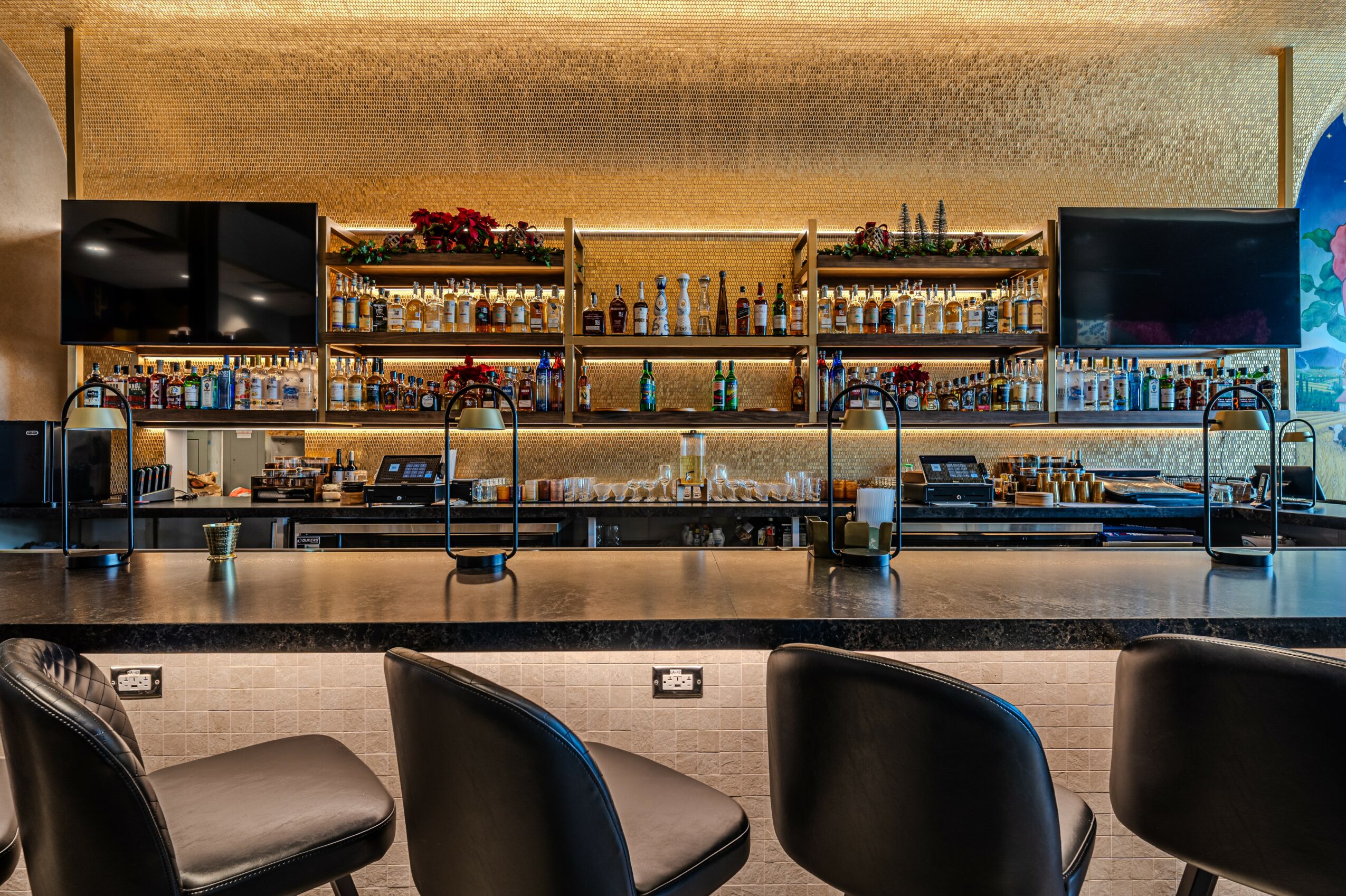 Bar counter with shelves of bottles, two monitors, and three chairs in front, in a modern setting.