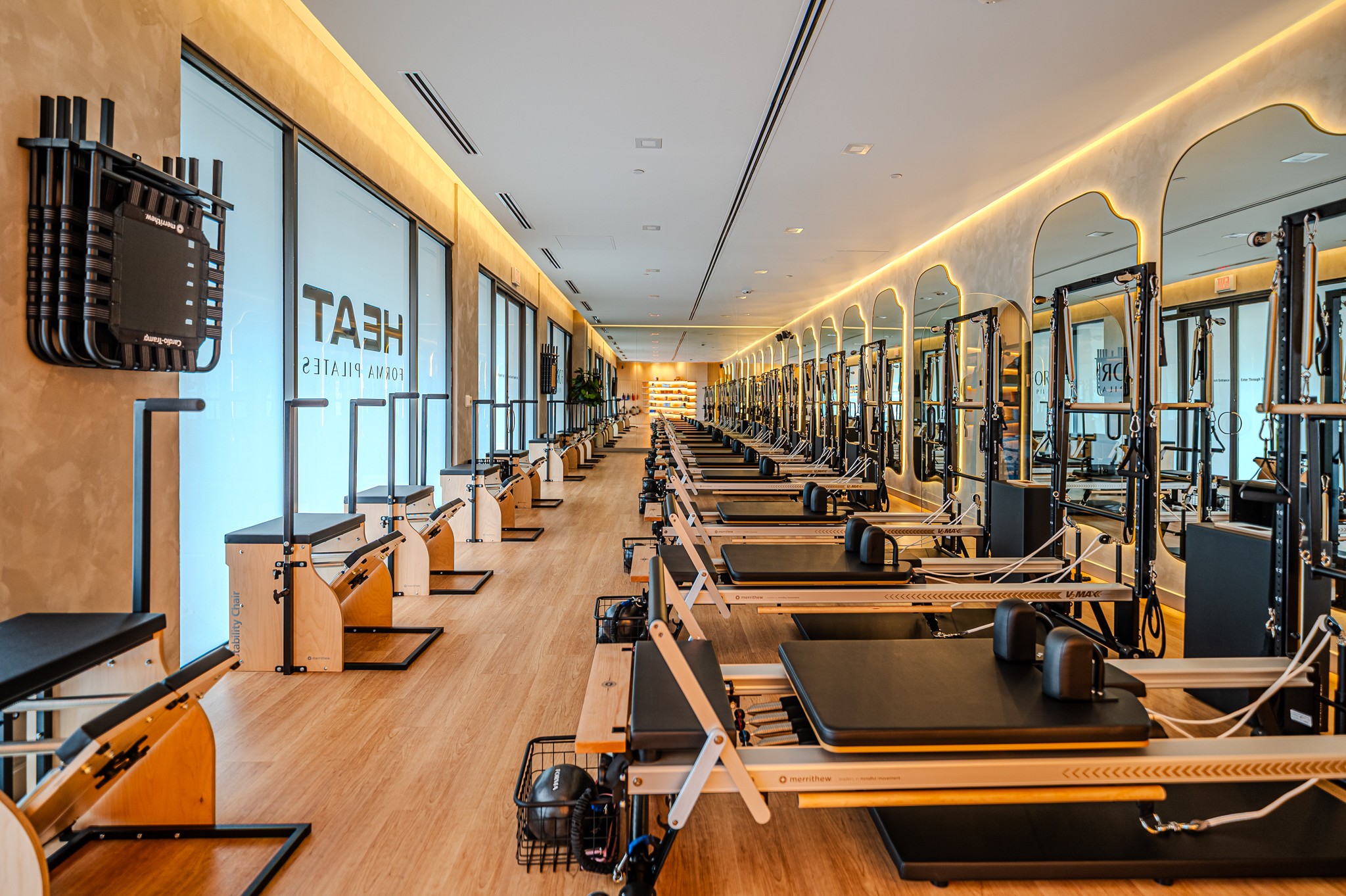 Empty fitness studio with rowing machines and mirrors, illuminated by warm lighting, with large windows on the right side.