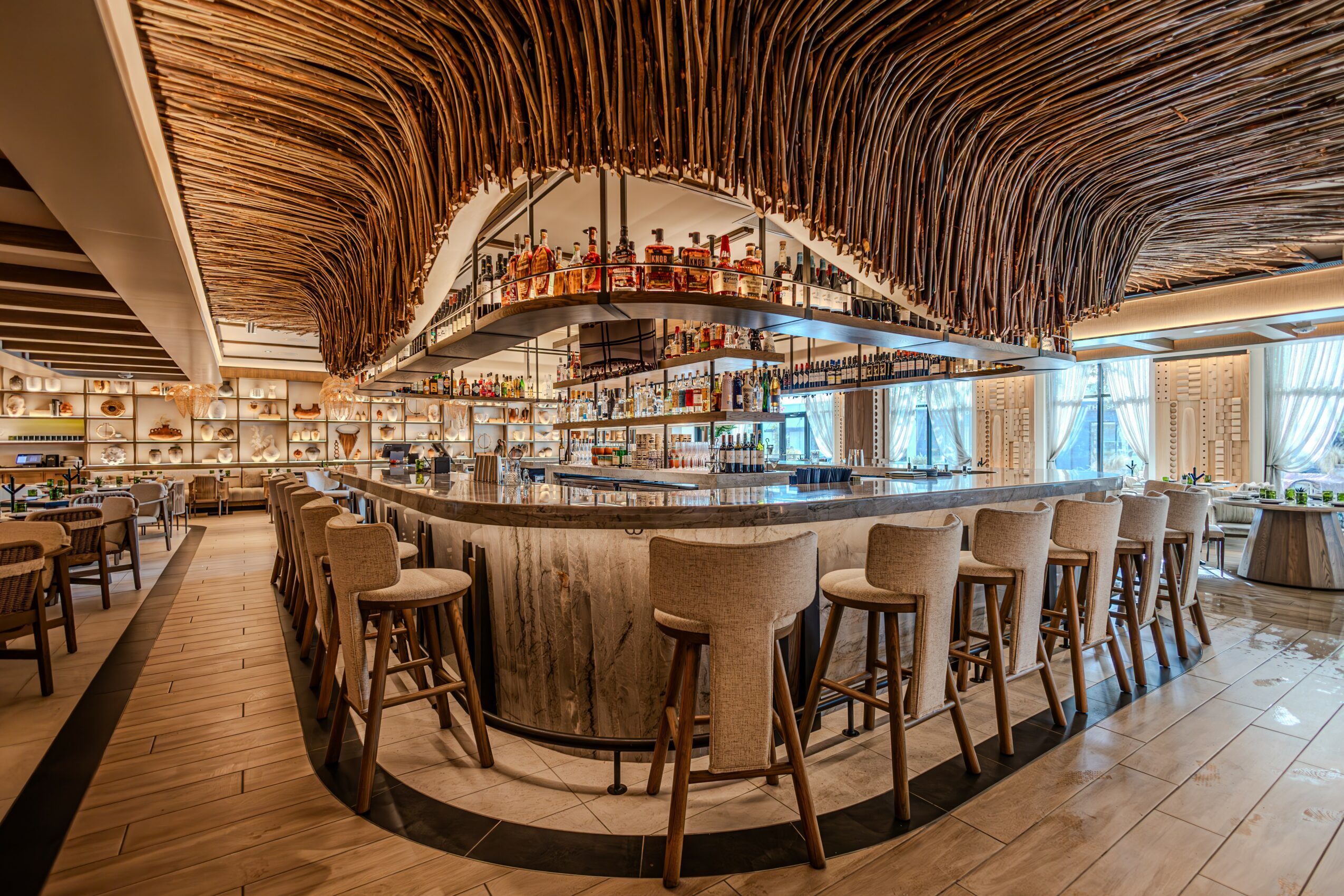 Bar with curved counter, high chairs, and a thatched ceiling design, with large windows and seating area in the background.