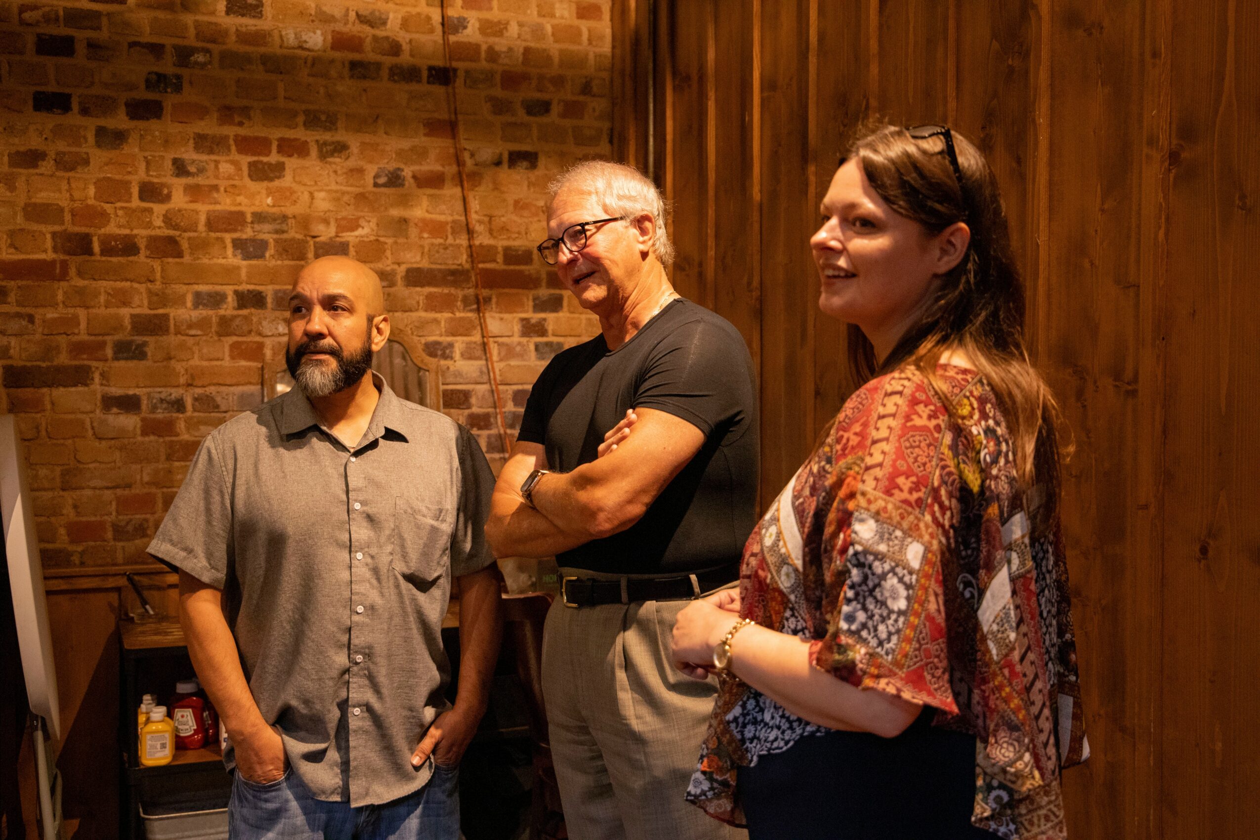 Three people standing and smiling indoors with brick and wooden walls.