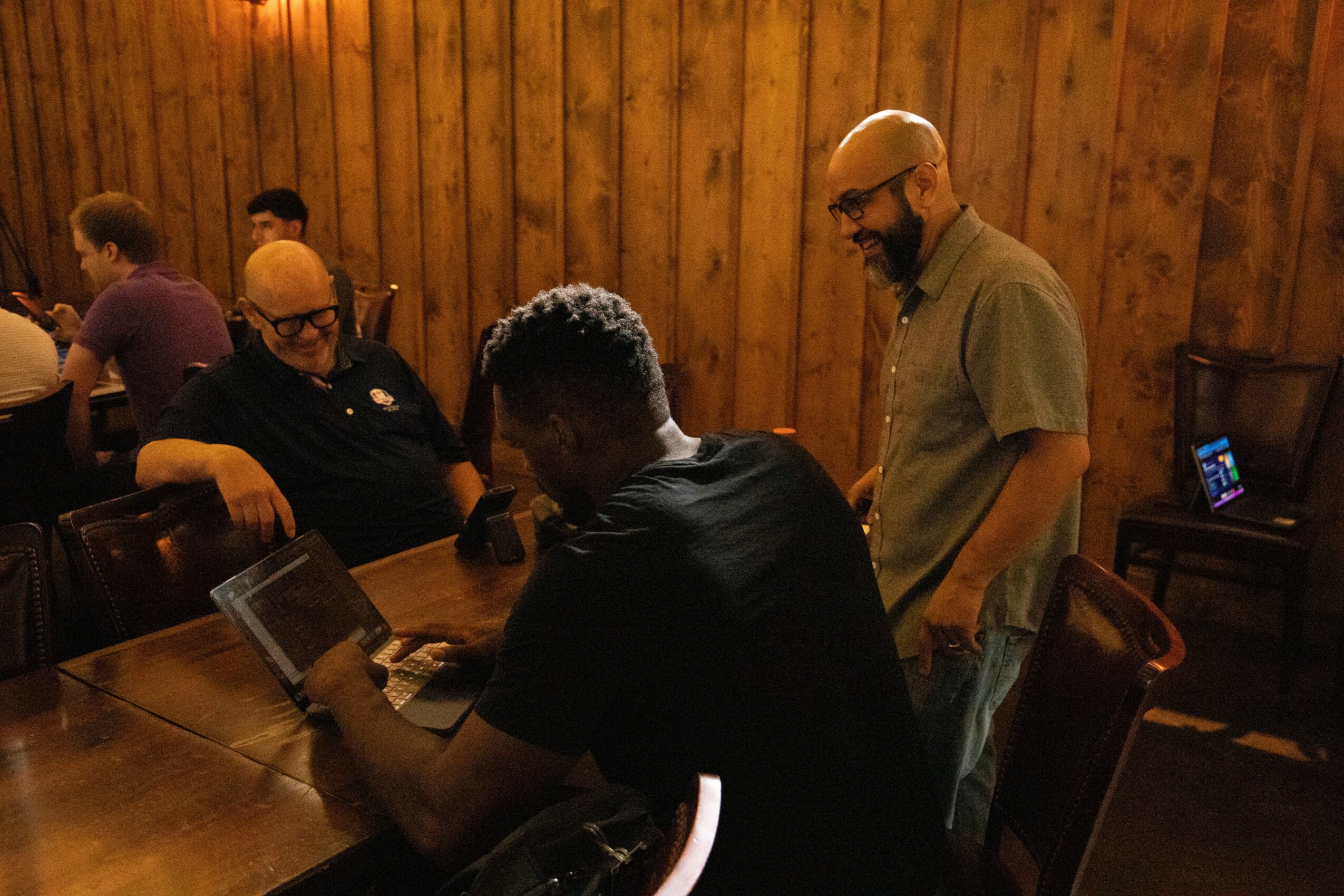 Three men sit and stand around a wooden table in a room with wood-paneled walls, using laptops and devices.