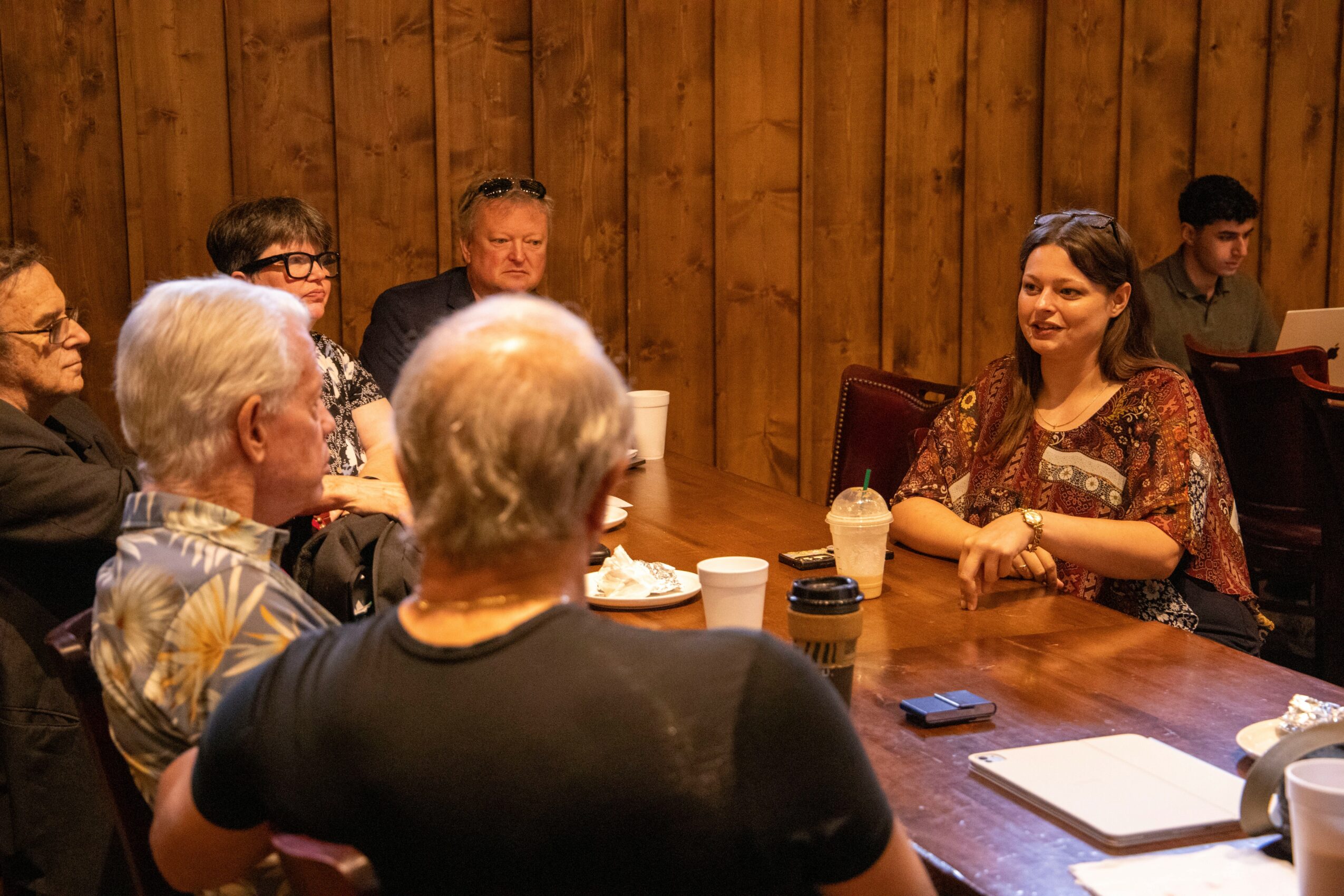 Group of people sitting around a wooden table in a room with wood-paneled walls, engaged in conversation, with drinks and notebooks on the table.