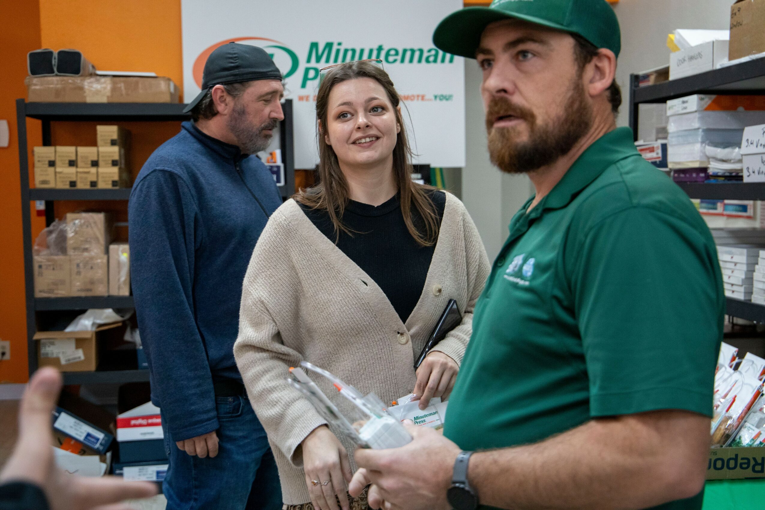 Three people in a store aisle, one man in green uniform holding a product, woman smiling, another man looking at shelves.