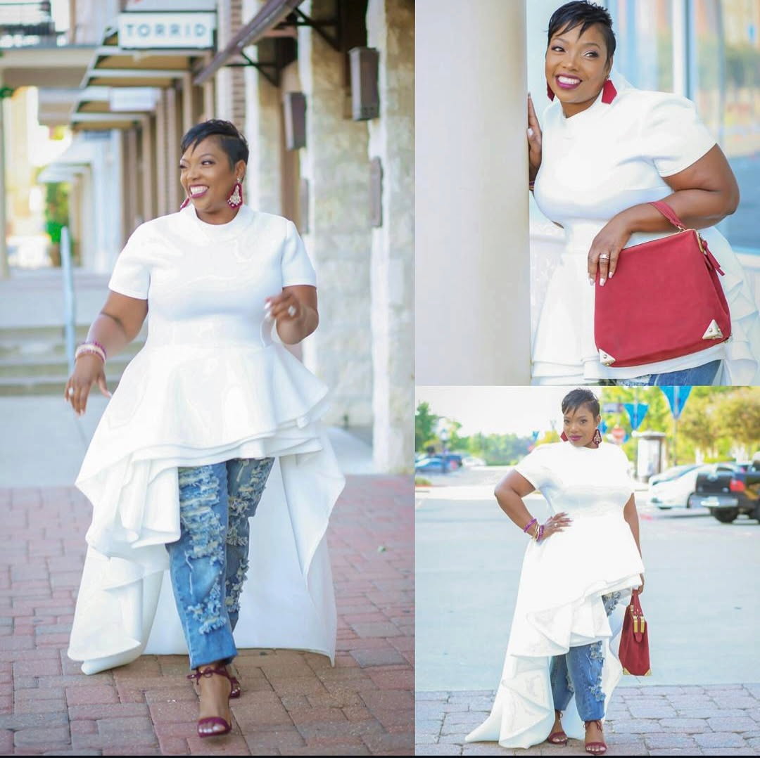 Woman in white dress and jeans walking on sidewalk, smiling, holding a red bag, in outdoor urban setting.