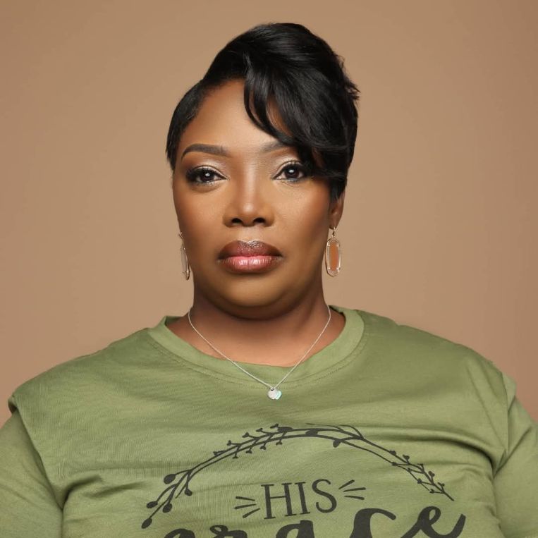 Portrait of a woman with short black hair, wearing a green shirt and jewelry, against a beige background.