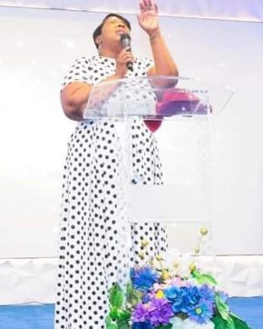 Woman speaking at a podium with a floral arrangement, wearing a polka dot dress, raising her hand, holding a microphone.