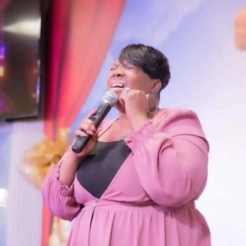 Person singing into a microphone with eyes closed, wearing a pink dress and black top, on stage with colorful curtains.