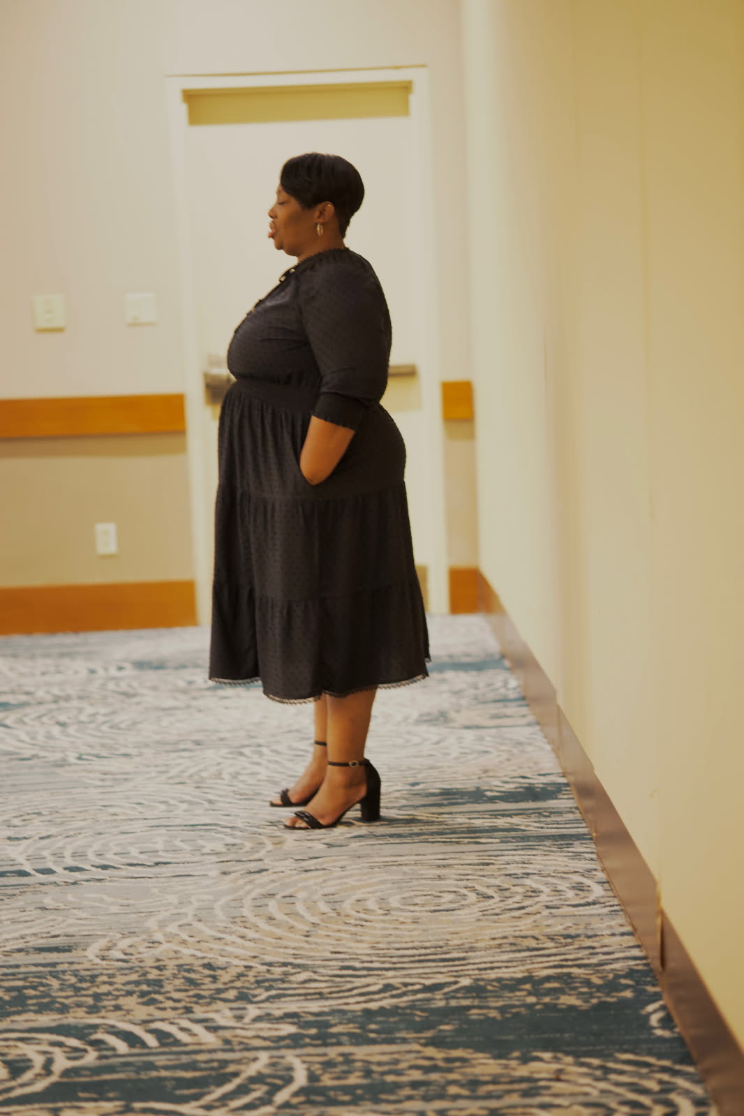 Woman standing in profile with hands in pockets, wearing a black dress and heels, in an indoor setting.