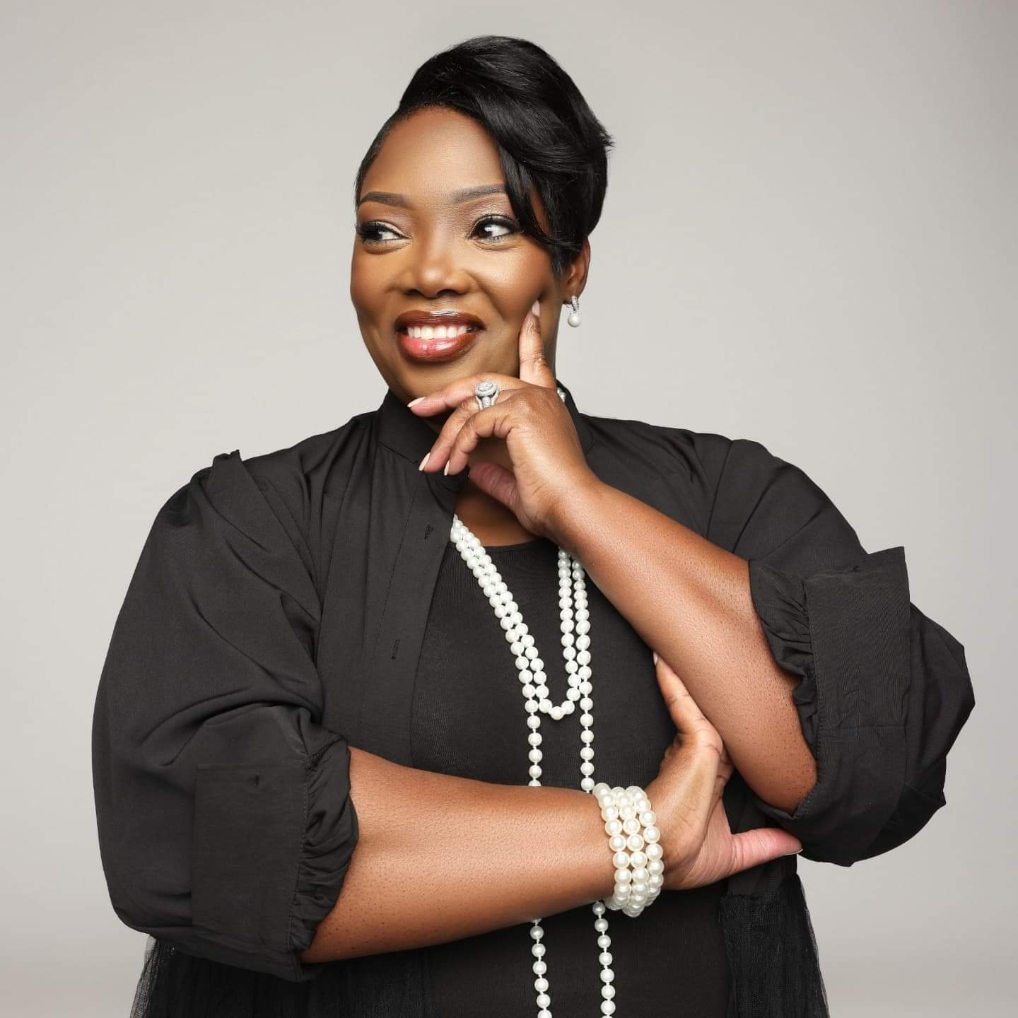 Woman with short black hair wearing a black dress and pearl jewelry, smiling and posing with her hand near her face.