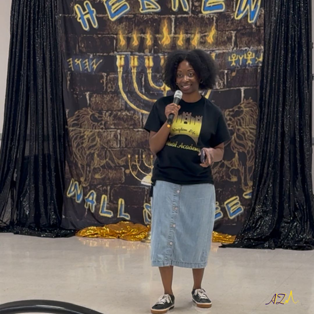 Woman standing on stage holding microphone, smiling, in front of a decorated backdrop with black curtains and neon signs.