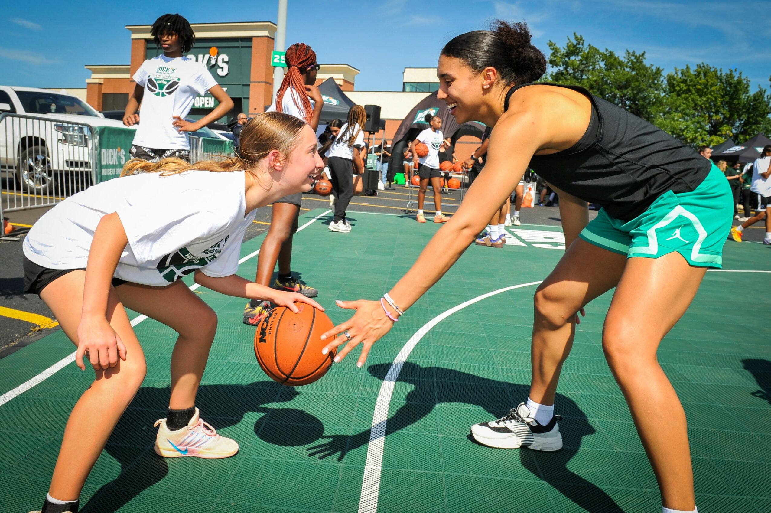 Young girl dribbling basketball while smiling at woman on outdoor court, with people and cars in background.