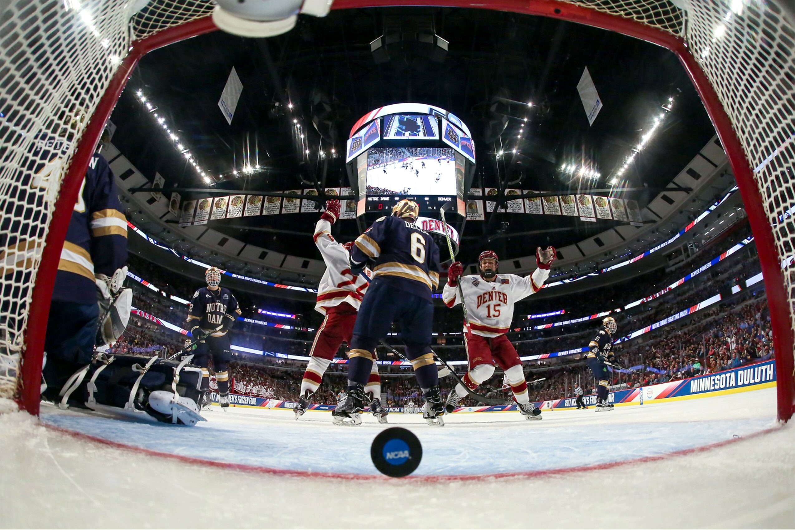 Hockey game scene with players near the goal, puck on ice, and scoreboard overhead in arena.