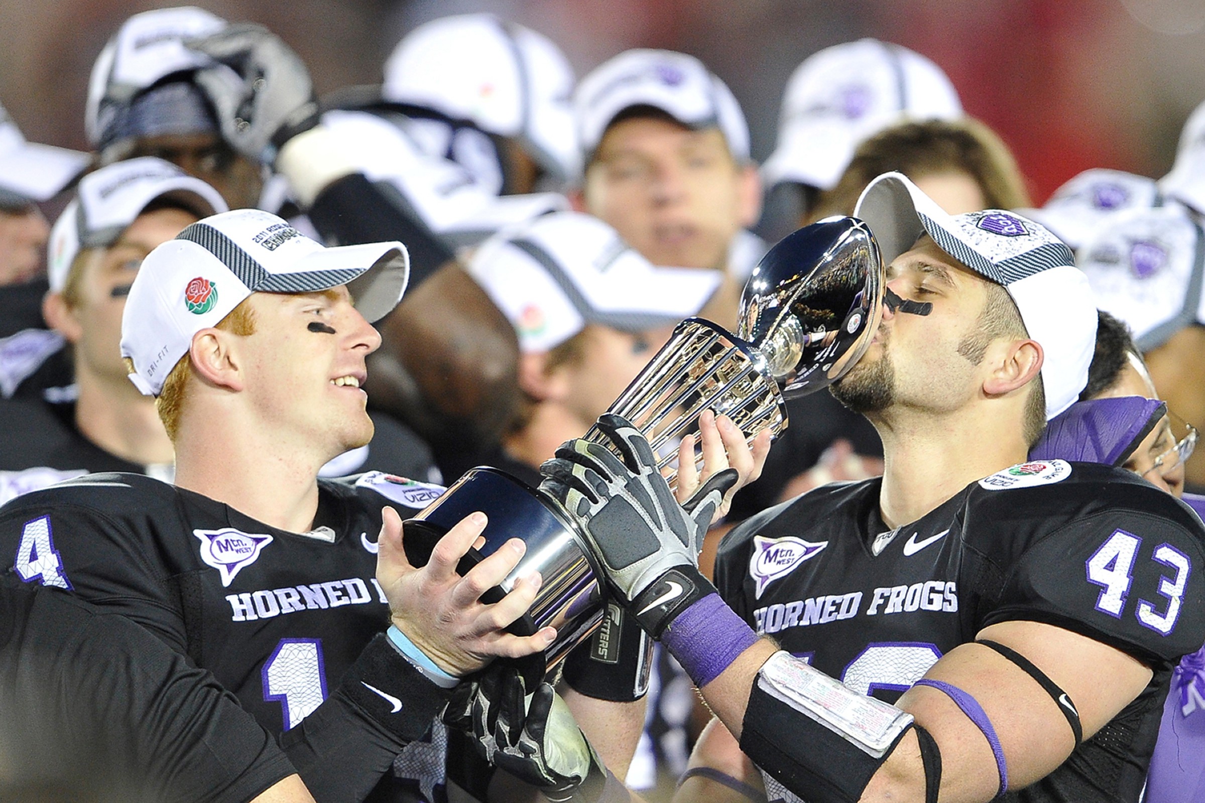Two football players in black uniforms and white caps share a trophy, smiling and kissing it, with teammates in background.