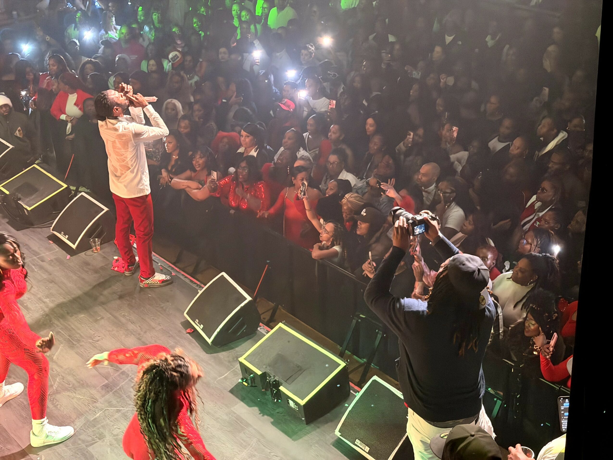 Performer on stage with microphone, audience watching and taking photos, stage monitors visible, lively event atmosphere.