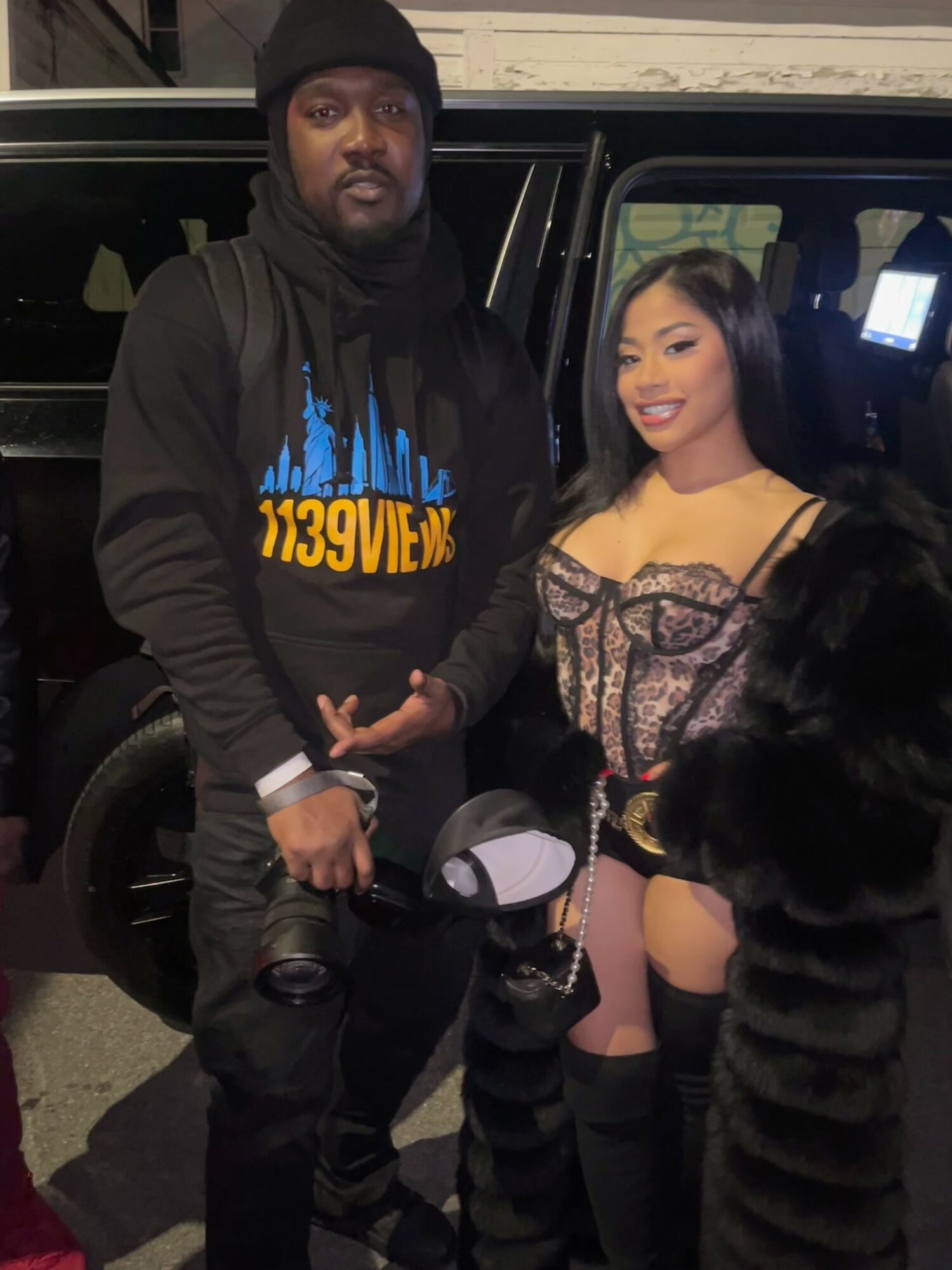 A man and woman stand together in front of a vehicle at night, smiling and posing for the photo.