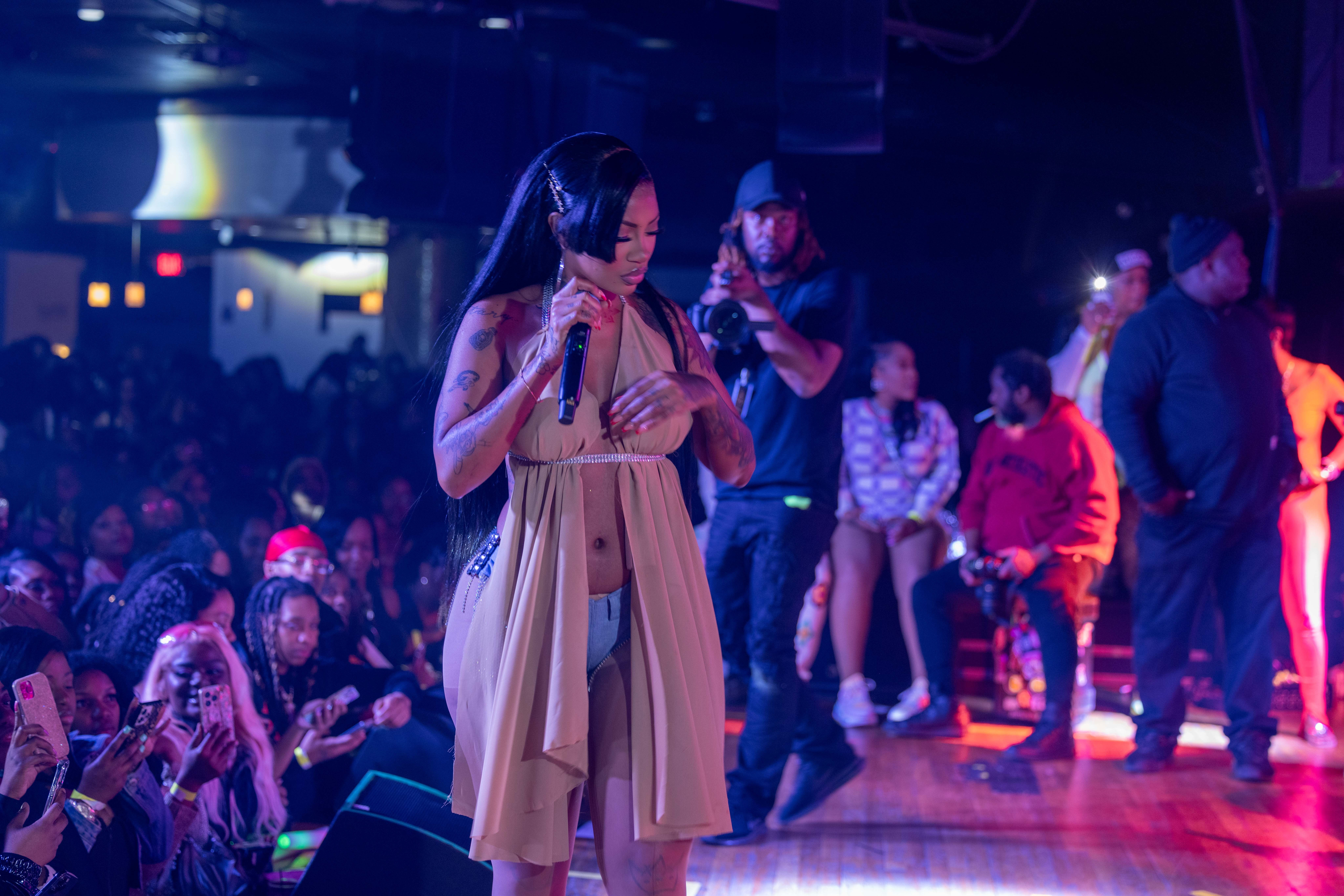 A woman in a beige dress on stage with a microphone, audience watching in a dimly lit venue.